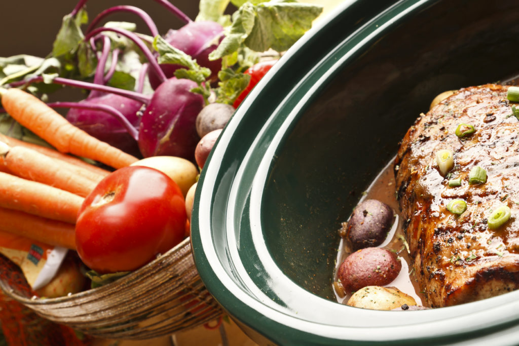 Slow cooker with pot roast and potatoes sitting next to a basket of fresh vegetables