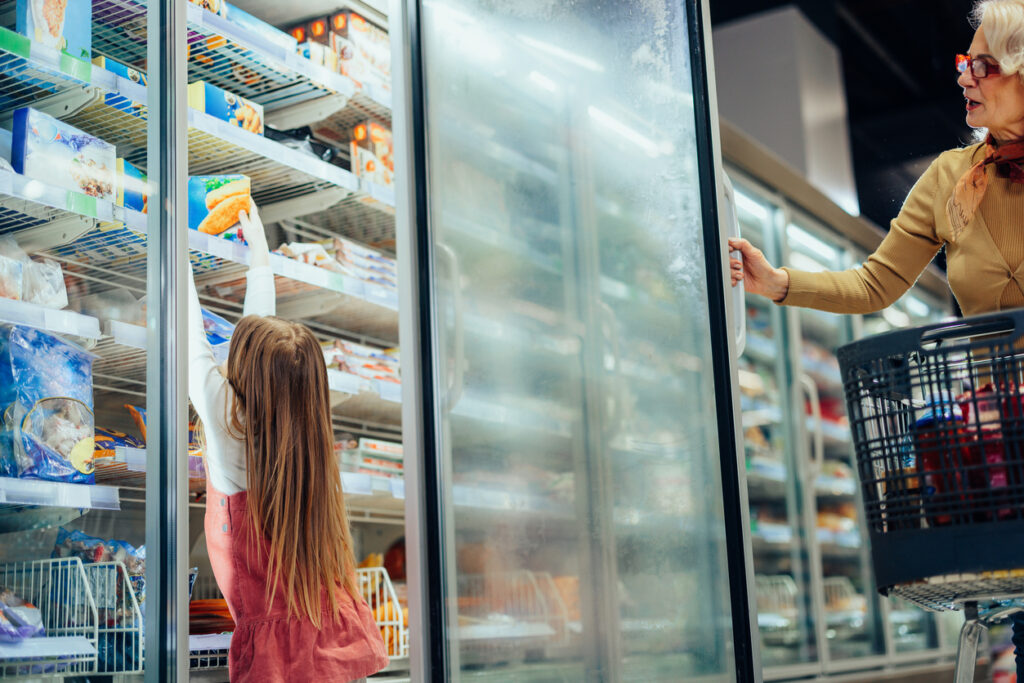 Young girl reaching for a box of frozen fish in the grocery freezer section while a shopper with a cart holds the door open