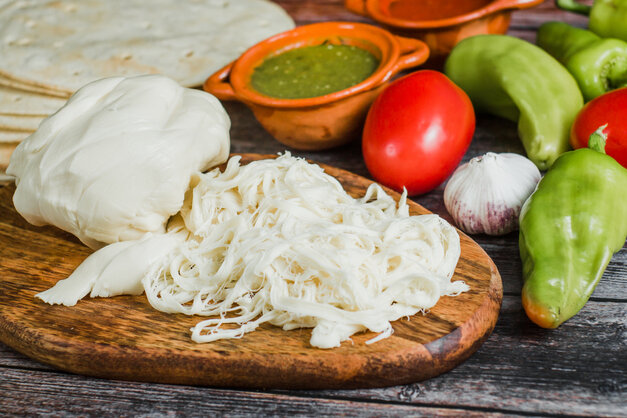 Mexican cheeses on a wooden platter next to peppers, tomatoes, garlic, salsa and tortillas